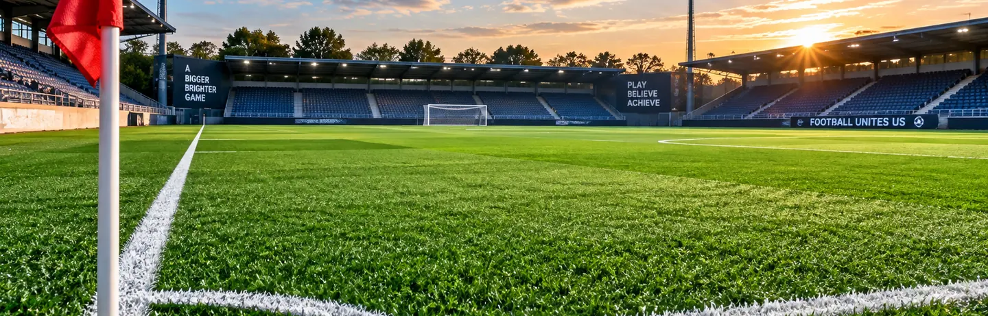 A football pitch at sunset, seen from beside the corner flag, with floodlights and empty stands behind the goal.